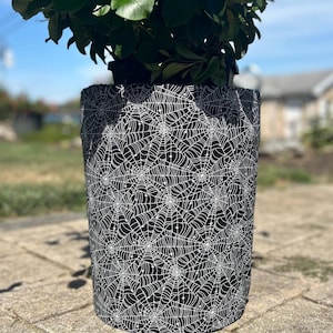 May include: A black and white spiderweb-patterned plant pot cover. The cover is cylindrical and surrounds a potted plant with green leaves. The design features a repeating pattern of white spiderwebs on a black background.
