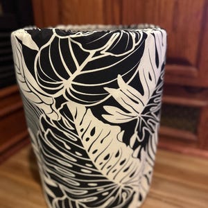 May include: A cylindrical fabric laundry hamper featuring a high-contrast black and white botanical print with large monstera leaves. The top edge is finished with a gathered elastic casing, creating a soft, rounded opening for the container.