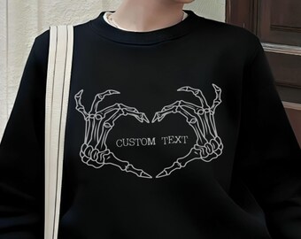 May include: Black sweatshirt with a white outline of two skeleton hands making a heart shape. The text "CUSTOM TEXT" is inside the heart.