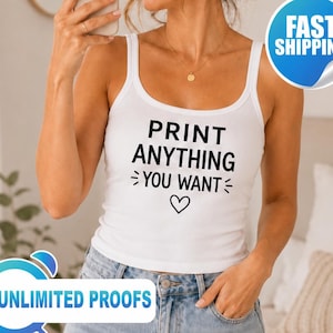 May include: A white tank top featuring the black text PRINT ANYTHING YOU WANT with decorative lines and a heart symbol centered on the chest. The garment has a scoop neckline and wide shoulder straps, worn with light blue denim jeans.