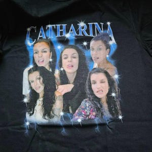 Pu&ograve; includere: Una t-shirt nera con una stampa grafica di quattro ritratti della stessa persona con lunghi capelli scuri. Il testo CATHARINA &egrave; stampato in un carattere blu metallizzato nella parte superiore, circondato da stelle e fulmini blu.