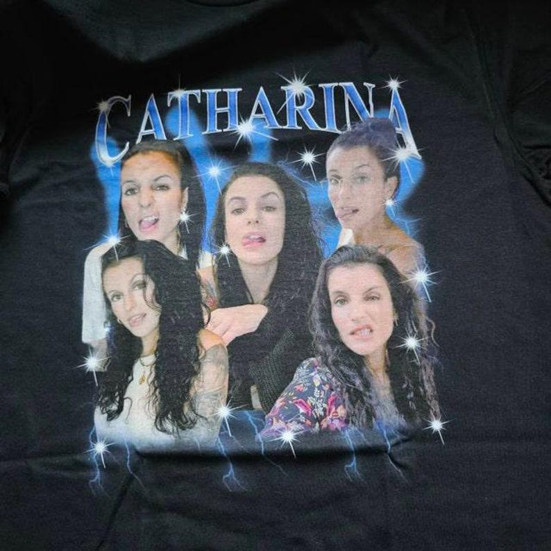 Pu&ograve; includere: Una t-shirt nera con una stampa grafica di quattro ritratti della stessa persona con lunghi capelli scuri. Il testo CATHARINA &egrave; stampato in un carattere blu metallizzato nella parte superiore, circondato da stelle e fulmini blu.