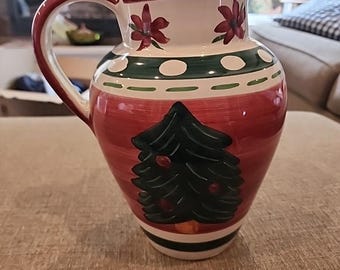 May include: A ceramic pitcher with a red base, green and white bands, and a Christmas tree design. The handle and rim are red, and the top features floral accents. A festive decorative piece.