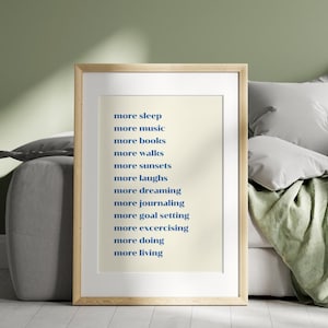 May include: A framed print with a cream background and blue text. The text lists aspirations including "more sleep," "more music," and "more living." The frame is light wood. The print is on a sofa with cushions and a blanket.