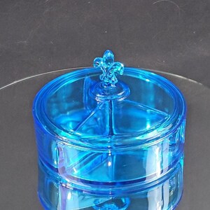 Könnte beinhalten: Eine blaue, geteilte Servierplatte aus Glas mit einem dekorativen Fleur-de-lis-Knopf auf dem Deckel.