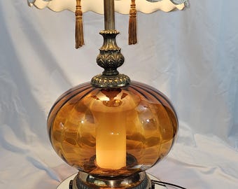 May include: A vintage amber glass table lamp with a white lampshade and gold accents. The lamp has a round base with a mirrored bottom.