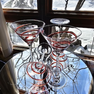 May include: Several clear martini glasses with red spiral detailing. The glasses are displayed on a mirrored surface, with a winter scene visible through a window in the background.