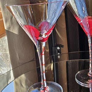 May include: Two clear martini glasses, each with red liquid. The glasses have a classic triangular bowl and long stems with red detailing. They sit on a reflective surface, with a neutral backdrop.