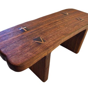 May include: A handcrafted wooden stool with a rich brown finish. The stool features a rectangular seat with decorative inlaid accents and two triangular legs. The seat is approximately 24 inches long.