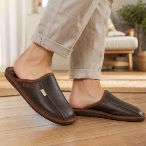 May include: Dark brown leather slippers with a textured surface and a contrasting brown trim. The slippers have a slip-on design and are shown on a wooden floor. The brand name "Nbasaco" is visible on the side of the slipper.
