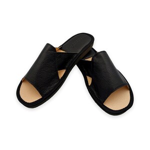 May include: A pair of black open-toe slide sandals featuring a textured leather upper with a wide strap design. The interior footbed is a light beige color, and the outer sole is black. The sandals have a simple, slip-on construction.