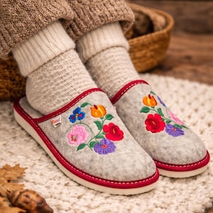 Puede incluir: Zapatillas de fieltro gris con diseños florales bordados en rojo, rosa, azul, naranja y morado. Las zapatillas tienen un ribete rojo y suelas blancas. La persona lleva calcetines beiges y pantalones marrones.