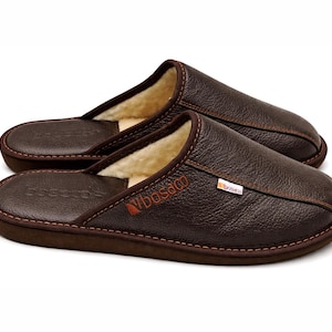 Pode incluir: Um par de chinelos de couro castanho com um forro macio de cor creme. Os chinelos têm um design costurado nas bordas e um pequeno logotipo na lateral. A marca "Vibes&co" é visível.