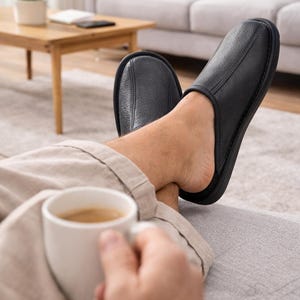 Puede incluir: Zapatillas de cuero negro con punta redondeada y detalles cosidos. Las zapatillas se usan en interiores. Una persona sostiene una taza de café blanca. El escenario es una sala de estar con un sofá y una mesa de centro.