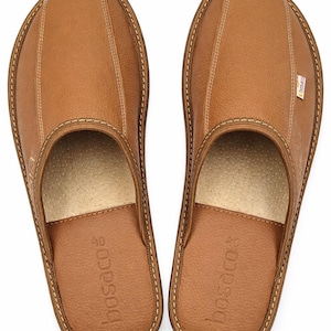 May include: A pair of brown leather slippers with a stitched design. The slippers have a rounded toe and a soft, cushioned insole. The brand name "bosaco" is printed on the side of each slipper. The slippers are designed for indoor use.