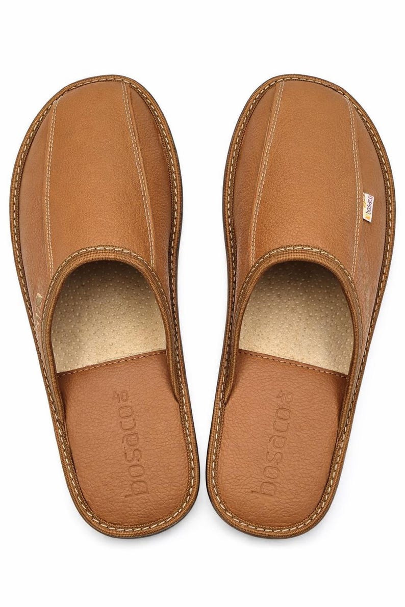 May include: A pair of brown leather slippers with a stitched design. The slippers have a rounded toe and a soft, cushioned insole. The brand name "bosaco" is printed on the side of each slipper. The slippers are designed for indoor use.