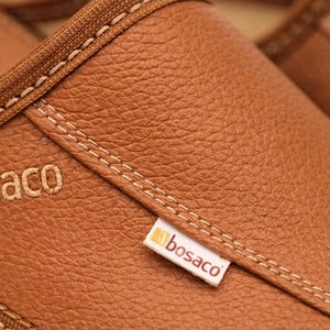May include: Close-up of brown leather footwear with visible stitching and the brand name "bosaco" embroidered on the side. The leather has a textured appearance, and the stitching is in a contrasting color.