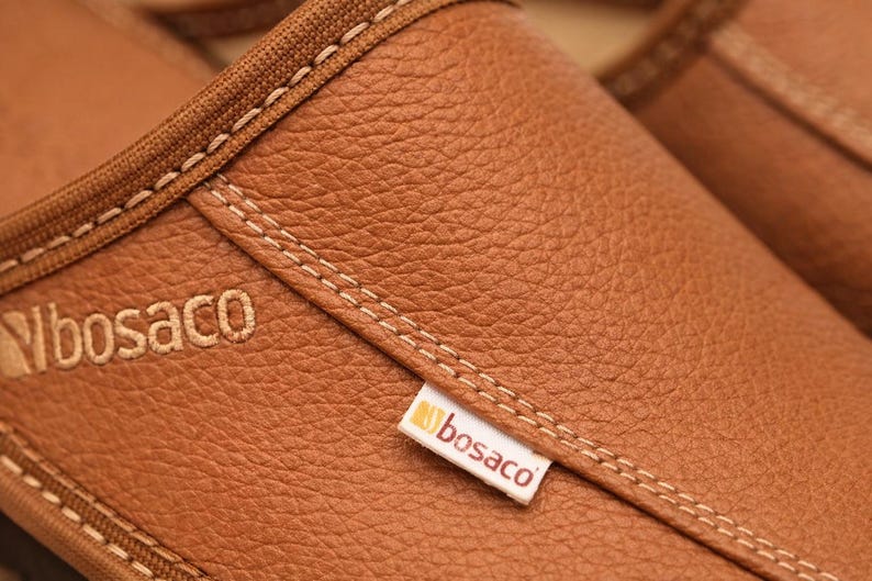 May include: Close-up of brown leather footwear with visible stitching and the brand name "bosaco" embroidered on the side. The leather has a textured appearance, and the stitching is in a contrasting color.