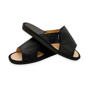 May include: A pair of black open-toe slide sandals featuring a textured leather upper with a small square logo patch on the side. The interior footbed is a light beige color, and the outer sole is black with a textured tread pattern for grip.