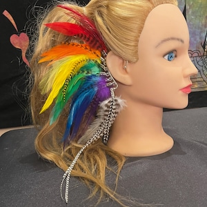 May include: A rainbow-colored feather ear cuff. The cuff features a spectrum of vibrant feathers, including red, orange, yellow, green, blue, and purple. Additional feathers are black and white. The ear cuff is attached to a silver-colored metal frame.