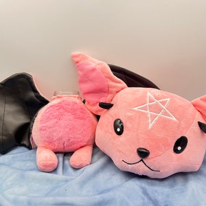 May include: Two pink plush toys shaped like bats with black wings. One bat has a white pentagram on its chest.