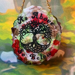 May include: A decorative resin pendant with a gold chain. The pendant features a black tree of life design with the words "As Above So Below" in black script. The background has a mix of red, green, brown, and white colors.