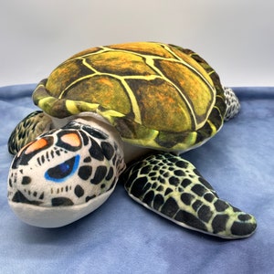 May include: A plush sea turtle toy with a green, brown, and black shell and a white face with black and orange markings. The turtle is lying on a blue fabric surface.