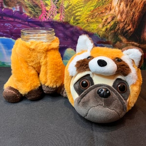 May include: A plush toy shaped like a pug wearing a red panda costume. The costume has a white face with black eyes and a black nose. The pug is wearing a brown and orange fur coat.