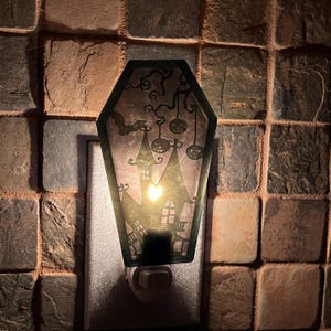 May include: A black and green nightlight shaped like a coffin with a silhouette of a haunted house, a bat, and pumpkins hanging from a tree. The nightlight is plugged into an outlet and is lit.