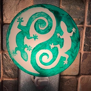 May include: Round night light with a teal and white design. The night light features two lizard silhouettes and a swirling pattern. The teal color has a marbled effect. The night light is plugged into a wall outlet.