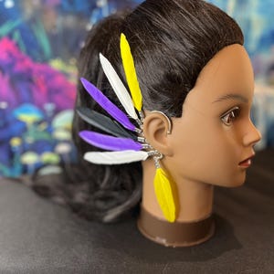 May include: A silver-toned ear cuff featuring a vertical arrangement of six feathers in yellow, white, purple, and black. The feathers are attached to a metal frame that contours the outer edge of the ear, with one yellow feather dangling below.