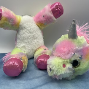 May include: A disassembled plush unicorn toy with a white body and colorful pastel accents. The unicorn's head and body are separate, with a silver horn, pink mane, and star-shaped embellishments. The hooves and ears are a shiny pink material.