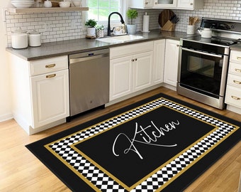 May include: A black kitchen floor mat with a gold border and a checkered pattern. The word "Kitchen" is written in a cursive font in the center of the mat. The mat is placed in front of a kitchen sink and dishwasher.