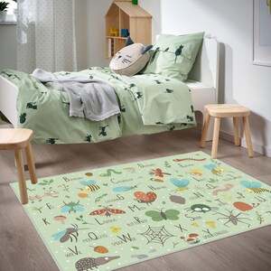 Op de afbeelding: Een lichtgroene kinderslaapkamer set met een bed, beddengoed en een vloerkleed. Het vloerkleed heeft een alfabet met insecten illustraties. Het beddengoed en de kussenslopen hebben een katten silhouet patroon. Een houten kruk en een klein houten huisje zijn ook zichtbaar.