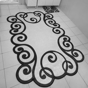 May include: A large, oval-shaped rug with a white center and a black, ornate border featuring swirling, scroll-like designs. The rug is placed on a tiled floor, and a smaller patterned mat is visible in the background. The rug's design adds a decorative element to the room.