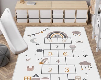 May include: A white hopscotch play mat with a whimsical design featuring a rainbow, clouds, and a rocket ship. The mat has numbered squares and decorative elements in neutral tones. A white slide is on the left.