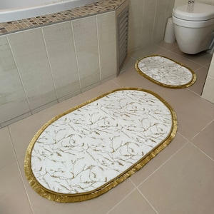 Peut inclure: Deux tapis de bain ovales avec un fond blanc et un motif de branches dorées. Les tapis ont une bordure en frange dorée.
