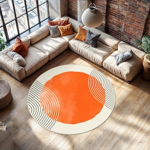 May include: A round area rug with an abstract design. The rug features a large orange watercolor-style circle in the center, surrounded by a cream-colored border. Black curved lines and stripes are on the sides. The rug is on a hardwood floor.