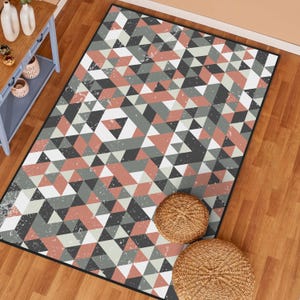 May include: Geometric patterned area rug featuring a repeating design of triangles in shades of gray, white, and coral. The rug has a black border and a distressed, vintage look. Two woven, round ottomans are placed on the wooden floor next to the rug.