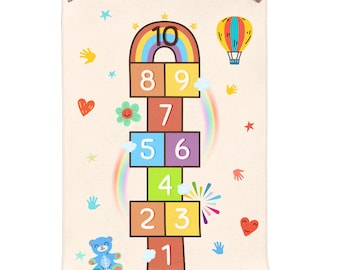 May include: A beige hopscotch play mat with colorful numbers, a rainbow, and a hot air balloon. The mat features a blue teddy bear, hearts, stars, and handprints. The numbers 1 through 10 are displayed.