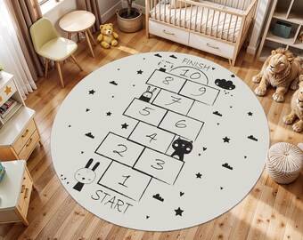 May include: A round, off-white rug with a black hopscotch design. The rug features a hopscotch grid with numbers, clouds, and star illustrations. The word "START" is at the beginning, and "FINISH" at the end. The rug is in a child's room.
