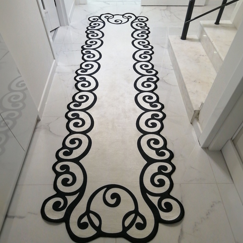 May include: A long, rectangular rug with a cream-colored center and a black, ornate scrollwork border. The rug is placed on a white marble floor, and the design is symmetrical, creating a decorative runner for a hallway or entryway.