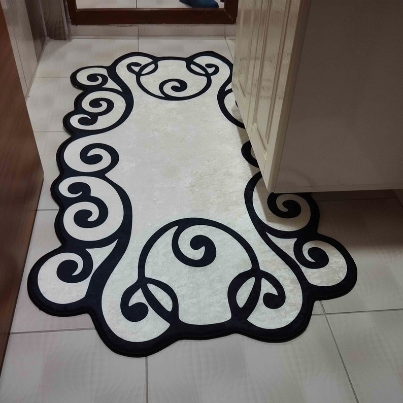 May include: A decorative rug with a cream-colored center and a black, swirling border. The rug has an oblong shape and is placed on a tiled floor. The design features a repeating pattern of stylized curls and loops.