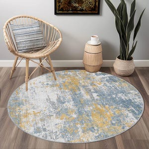 May include: A round area rug with a distressed design. The rug features a blend of white, blue, and yellow colors, creating a textured, abstract pattern. The rug is placed on a wooden floor, and is near a wicker chair.