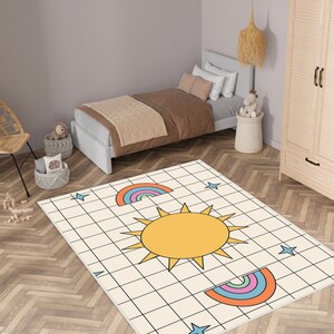 May include: A white rug with a grid pattern features a yellow sun with orange rays and two rainbow arches. The rug is decorated with blue stars.