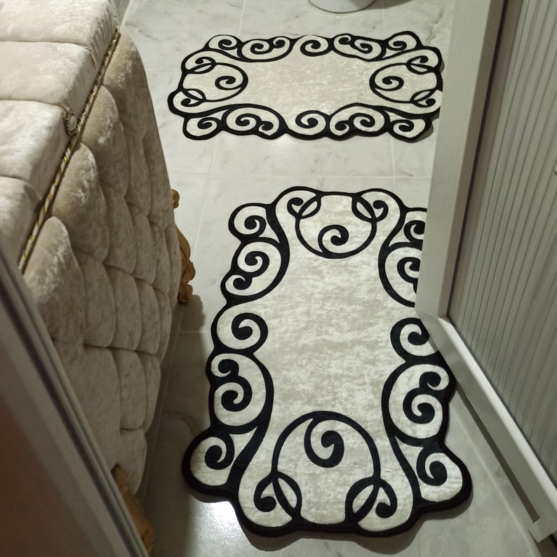 May include: Two ivory-colored bathroom rugs with a black, ornate, scrollwork border. One rug is rectangular, and the other is oval-shaped. The rugs are placed on a tiled floor near a beige upholstered bench.