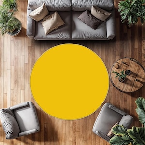 May include: A large, solid, circular rug in a vibrant mustard yellow color. The rug is positioned centrally on a wooden floor, surrounded by a gray sofa, two gray armchairs, a round wooden coffee table, and several potted green plants.