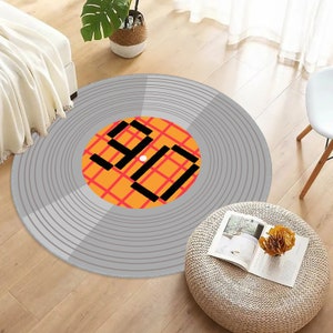 Könnte beinhalten: Ein runder Teppich mit einem grauen Schallplatten-Design und konzentrischen Rillen. Das mittlere Etikett ist orange mit einem roten Gittermuster und dem schwarzen, blockartigen Text 90, der deutlich in der Mitte der runden Fläche aufgedruckt ist.