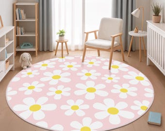 May include: A large, circular area rug featuring a repeating pattern of white daisy flowers with bright yellow centres on a soft pink background. The rug is positioned on a light-coloured wooden floor in a room with a chair, shelves, and a lamp.