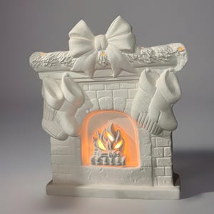 May include: A white ceramic Christmas fireplace ornament featuring a bow, garland, and two hanging stockings. The fireplace has an orange light inside, simulating a fire. A festive holiday decoration.